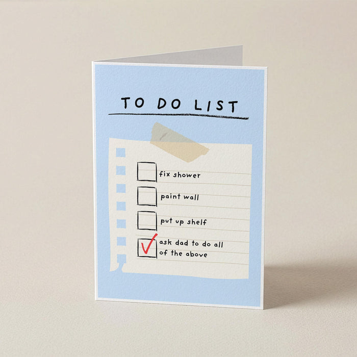 A folded greeting card standing upright on a light beige surface. The card has a pale blue border and reads 'TO DO LIST' above a checklist with a tape graphic and a red checkmark on the last item.