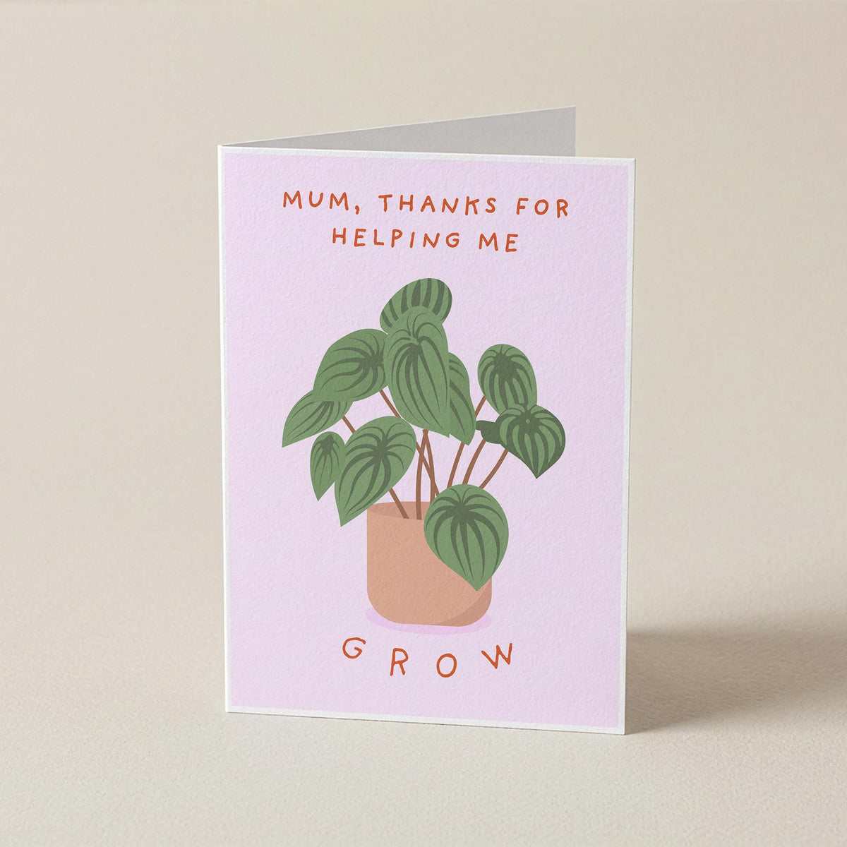 A folded greeting card standing upright on a light beige surface. The card has a pale pink background with an illustration of a green potted plant and the text 'MUM, THANKS FOR HELPING ME' at the top and 'GROW' at the bottom.