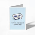 A folded greeting card shown standing upright on a white background. The card features a light blue center with an illustration of a Lurpak tub and the text 'I GOT YOU AN EXPENSIVE GIFT THIS YEAR!'.
