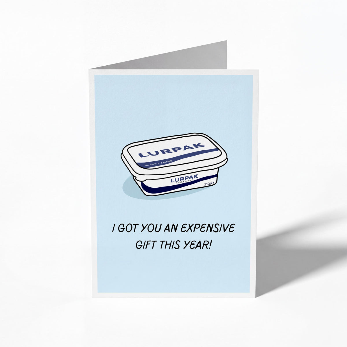 A folded greeting card shown standing upright on a white background. The card features a light blue center with an illustration of a Lurpak tub and the text 'I GOT YOU AN EXPENSIVE GIFT THIS YEAR!'.