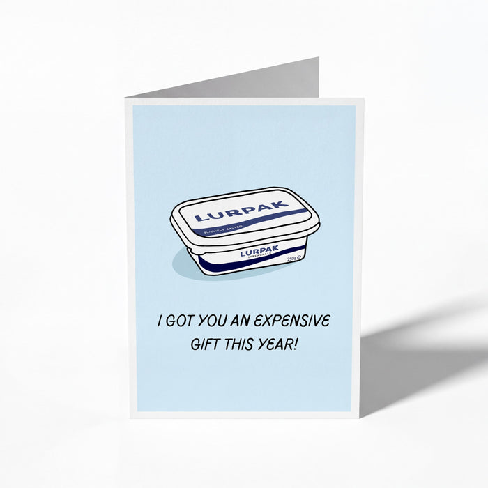A folded greeting card shown standing upright on a white background. The card features a light blue center with an illustration of a Lurpak tub and the text 'I GOT YOU AN EXPENSIVE GIFT THIS YEAR!'.