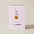 A folded greeting card standing upright on a light beige surface. The card has a pale pink front with a cartoon ghost wearing a party hat and holding a pumpkin, plus the text 'HAPPY BIRTHDAY, PUMPKIN!'.