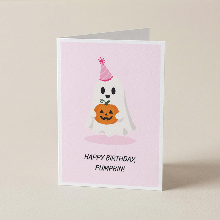 A folded greeting card standing upright on a light beige surface. The card has a pale pink front with a cartoon ghost wearing a party hat and holding a pumpkin, plus the text 'HAPPY BIRTHDAY, PUMPKIN!'.