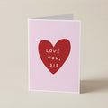 A folded greeting card standing upright on a light beige surface. The card has a pale pink background with a large red heart in the center containing the white text 'LOVE YOU, SIS'.