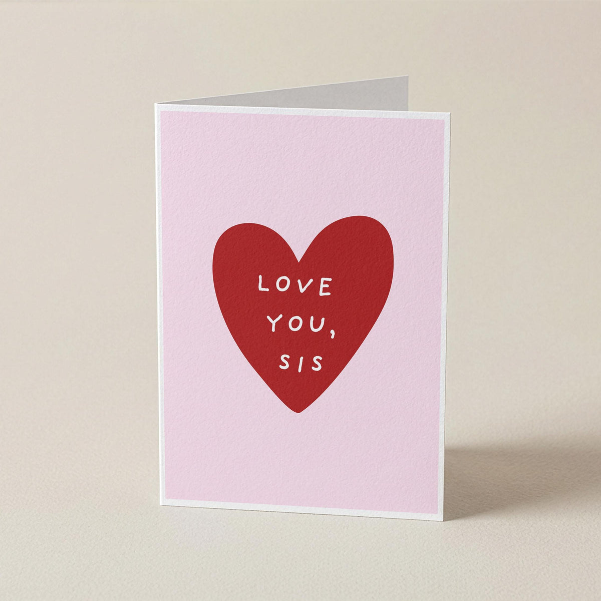 A folded greeting card standing upright on a light beige surface. The card has a pale pink background with a large red heart in the center containing the white text 'LOVE YOU, SIS'.