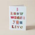 A folded greeting card standing upright on a beige surface. The card has a white background with the phrase 'I know where you live' printed in colorful, patchwork-style letters.