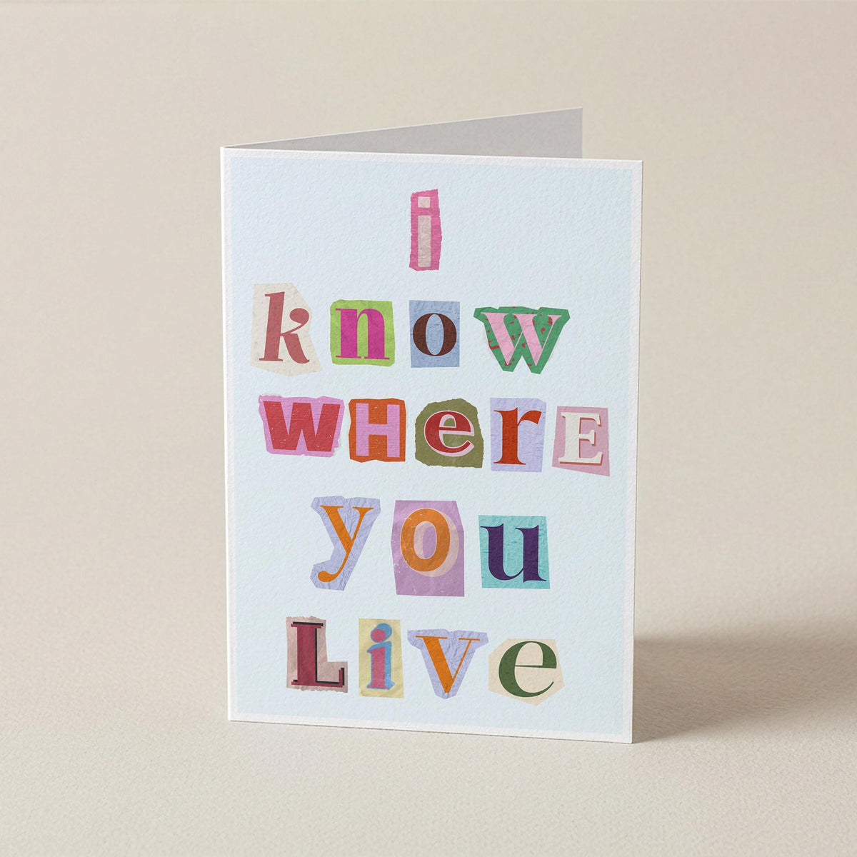 A folded greeting card standing upright on a beige surface. The card has a white background with the phrase 'I know where you live' printed in colorful, patchwork-style letters.
