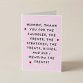 A folded greeting card standing upright on a light beige surface. The card is pale pink with black handwritten-style text reading, 'Mummy, thank you for the snuggles, the treats, the scratches, the treats, kisses, and did i mention the treats?' and small red heart accents.