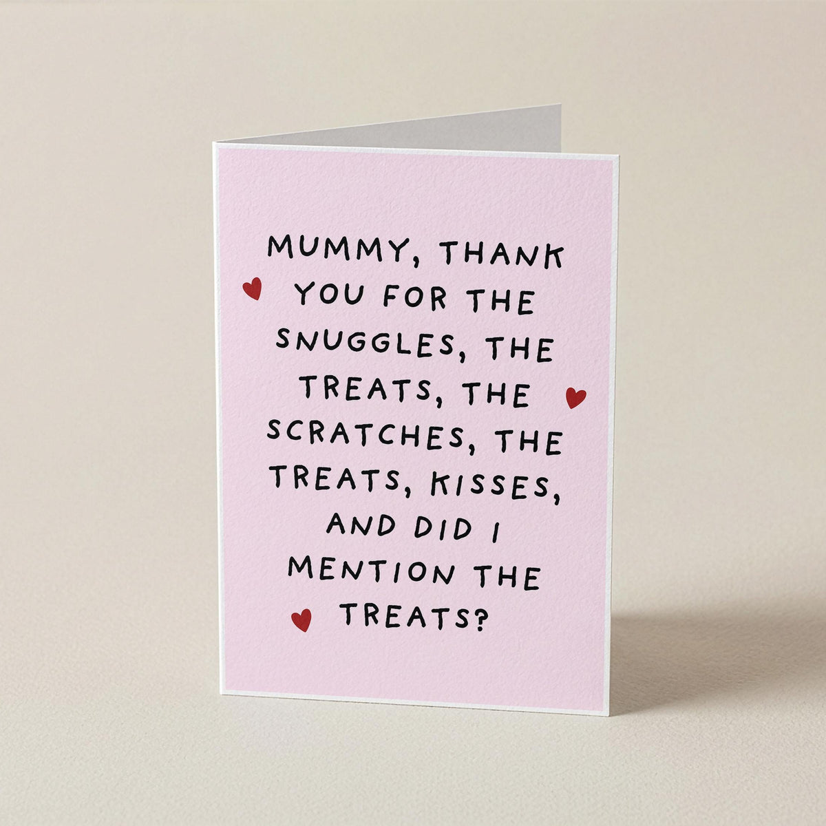 A folded greeting card standing upright on a light beige surface. The card is pale pink with black handwritten-style text reading, 'Mummy, thank you for the snuggles, the treats, the scratches, the treats, kisses, and did i mention the treats?' and small red heart accents.