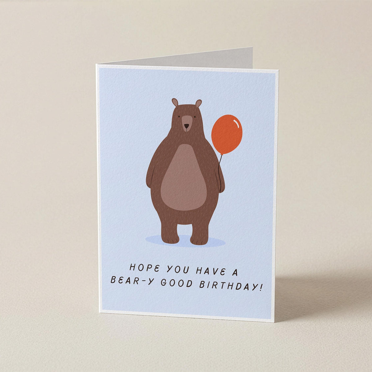 A folded greeting card standing upright on a light beige surface. The card features a light blue background with an illustration of a brown bear holding an orange balloon and the text 'HOPE YOU HAVE A BEAR-Y GOOD BIRTHDAY!'.