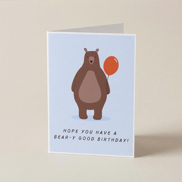 A folded greeting card standing upright on a light beige surface. The card features a light blue background with an illustration of a brown bear holding an orange balloon and the text 'HOPE YOU HAVE A BEAR-Y GOOD BIRTHDAY!'.