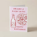 A folded greeting card standing upright on a light beige background. The card has a pale pink front with red line drawings of a bottle and a pizza slice, plus the text 'DRINKS & PICKY BITS! HAPPY BIRTHDAY!'.