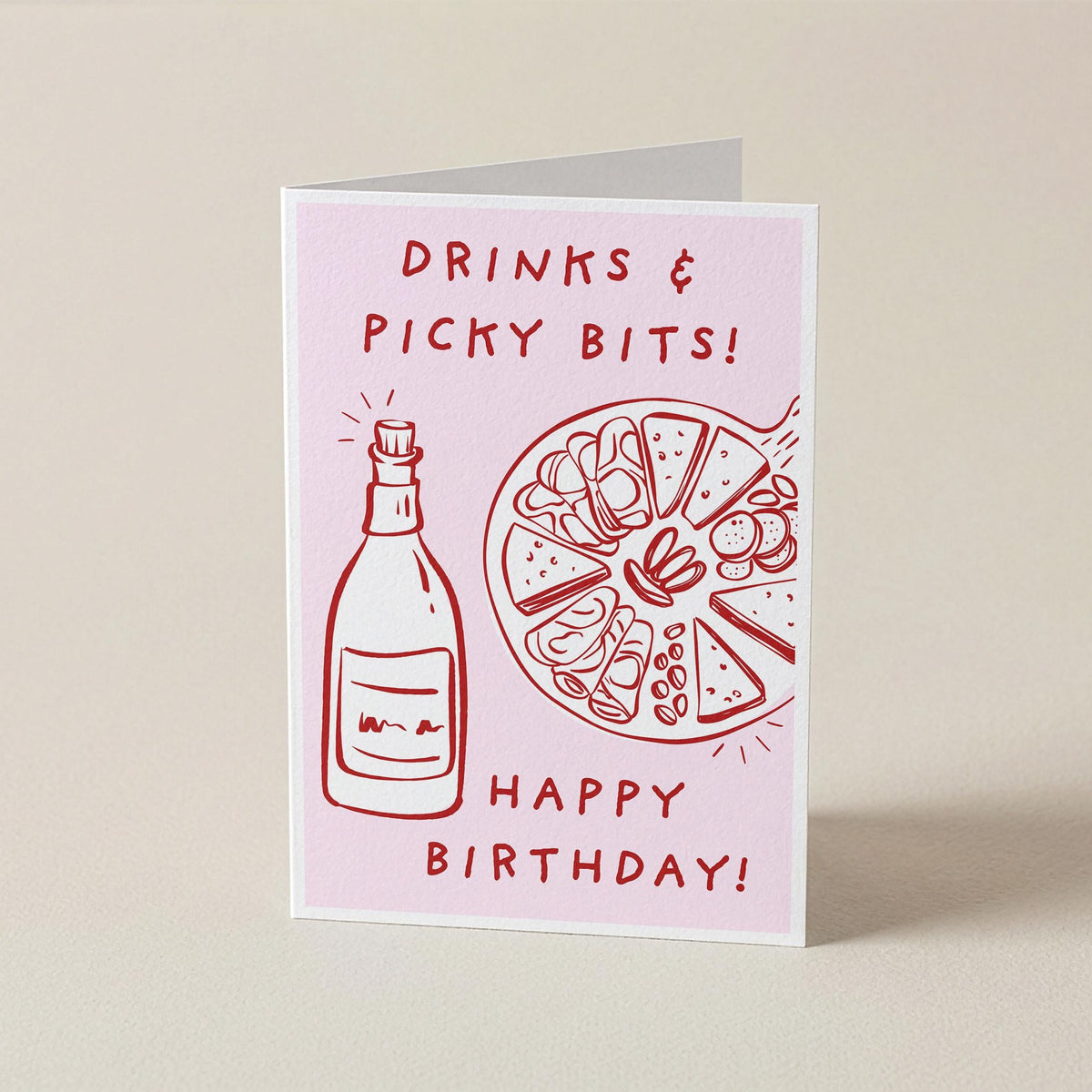 A folded greeting card standing upright on a light beige background. The card has a pale pink front with red line drawings of a bottle and a pizza slice, plus the text 'DRINKS & PICKY BITS! HAPPY BIRTHDAY!'.