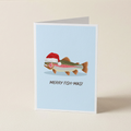 A folded greeting card standing upright on a light beige surface. The card has a pale blue background and features an illustrated fish wearing a red Santa hat with the text 'MERRY FISH-MAS!' below it.