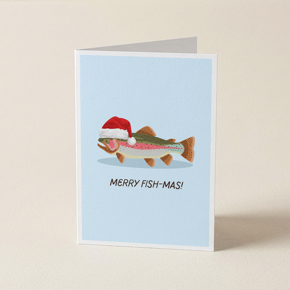 A folded greeting card standing upright on a light beige surface. The card has a pale blue background and features an illustrated fish wearing a red Santa hat with the text 'MERRY FISH-MAS!' below it.