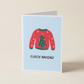 A folded greeting card standing upright on a light beige surface. The card has a pale blue background and features an illustration of a red Christmas sweater with a green tree on the front, plus the text 'FLEECE NAVIDAD' below.