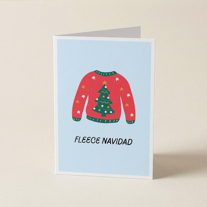 A folded greeting card standing upright on a light beige surface. The card has a pale blue background and features an illustration of a red Christmas sweater with a green tree on the front, plus the text 'FLEECE NAVIDAD' below.