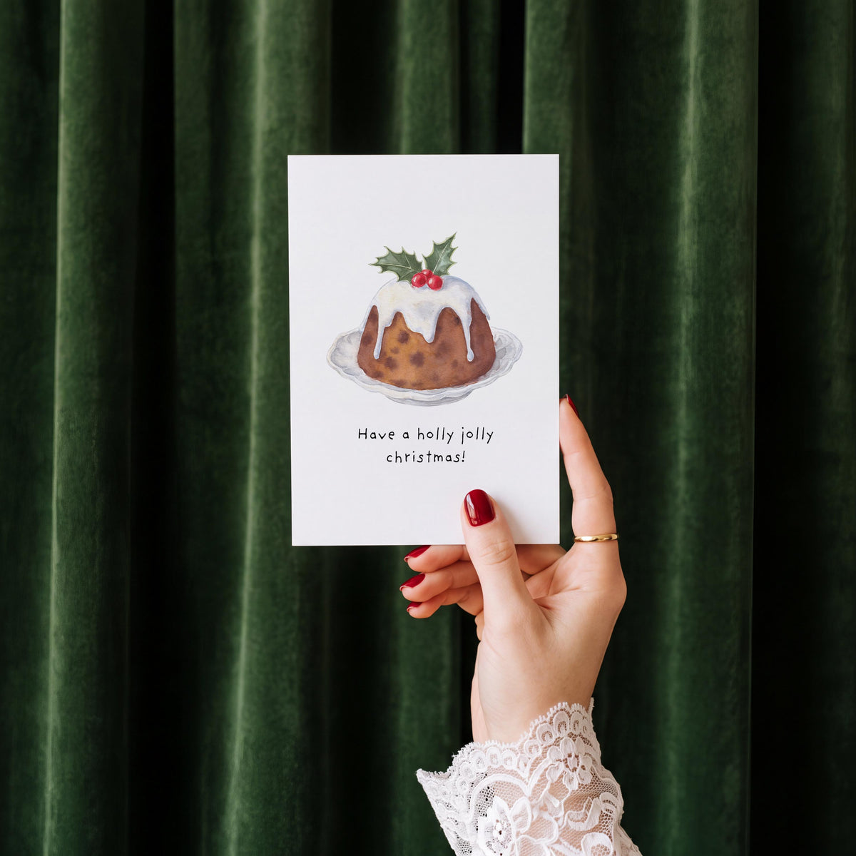 A hand with red nail polish and a white lace cuff holds a white greeting card featuring a watercolor illustration of a Christmas pudding with the text 'Have a holly jolly christmas!'. The background is a dark green velvet curtain.