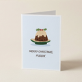 A folded Christmas greeting card standing upright on a light beige surface. The card has a pale blue front with an illustration of a christmas pudding on a plate and the text 'MERRY CHRISTMAS, PUDDIN'.