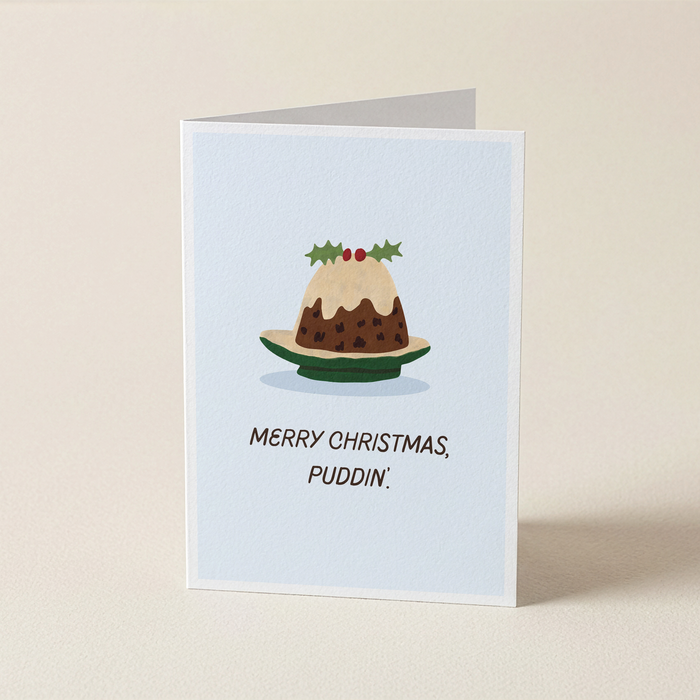 A folded Christmas greeting card standing upright on a light beige surface. The card has a pale blue front with an illustration of a christmas pudding on a plate and the text 'MERRY CHRISTMAS, PUDDIN'.