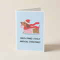 A folded greeting card standing upright on a light beige surface. The card has a pale blue front with an illustration of a brown dachshund wearing a red Santa hat and scarf, plus the text 'HAVE A PAWS-ITIVELY MAGICAL CHRISTMAS!'.