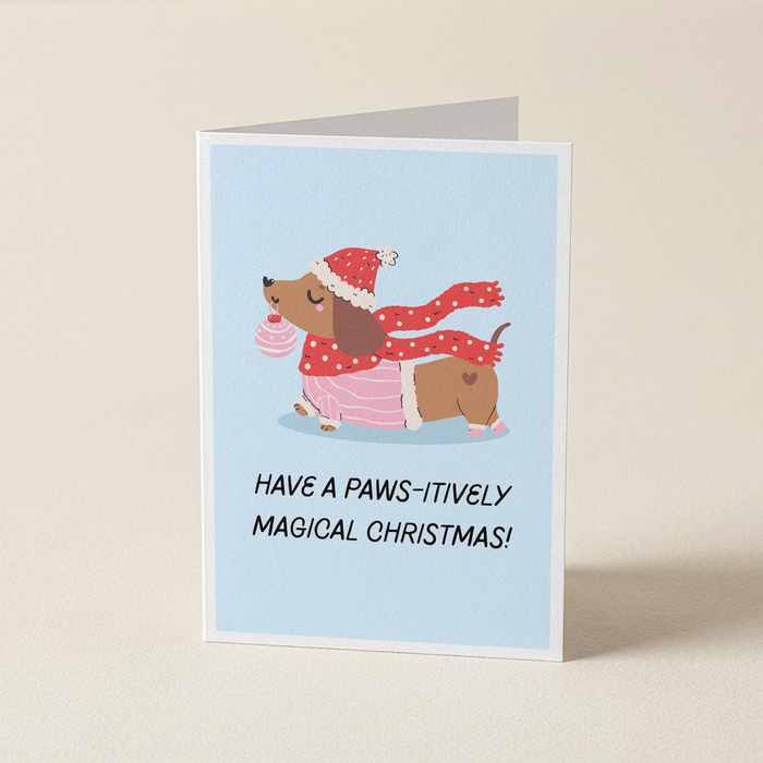 A folded greeting card standing upright on a light beige surface. The card has a pale blue front with an illustration of a brown dachshund wearing a red Santa hat and scarf, plus the text 'HAVE A PAWS-ITIVELY MAGICAL CHRISTMAS!'.