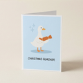 A folded greeting card standing upright on a light beige surface. The card features a white duck with orange legs and a christmas cracker in its hands, set against a pale blue background with the text 'CHRISTMAS QUACKER.