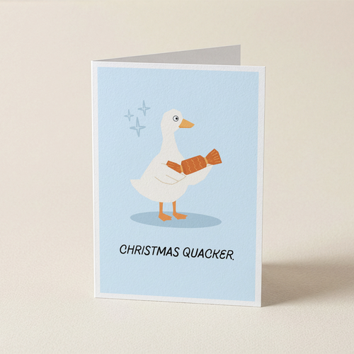 A folded greeting card standing upright on a light beige surface. The card features a white duck with orange legs and a christmas cracker in its hands, set against a pale blue background with the text 'CHRISTMAS QUACKER.