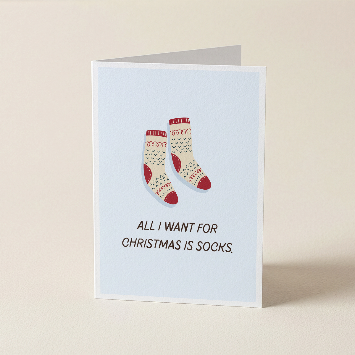 A folded greeting card standing upright on a light beige surface. The card has a pale blue front with an illustration of a pair of cream Christmas socks with red and green patterns, above the text 'ALL I WANT FOR CHRISTMAS IS SOCKS.