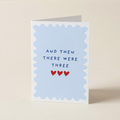 A folded greeting card standing upright on a light beige surface. The front has a pale blue scalloped border with the text 'AND THEN THERE WERE THREE' in dark blue letters and three small red hearts below.