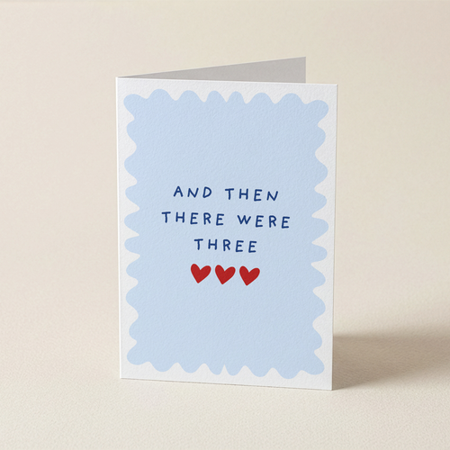 A folded greeting card standing upright on a light beige surface. The front has a pale blue scalloped border with the text 'AND THEN THERE WERE THREE' in dark blue letters and three small red hearts below.
