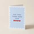 A folded greeting card standing upright on a light beige surface. The card has a white border with a scalloped light blue inner frame and the printed text 'AND THEN THERE WERE FOUR' above four small red hearts.