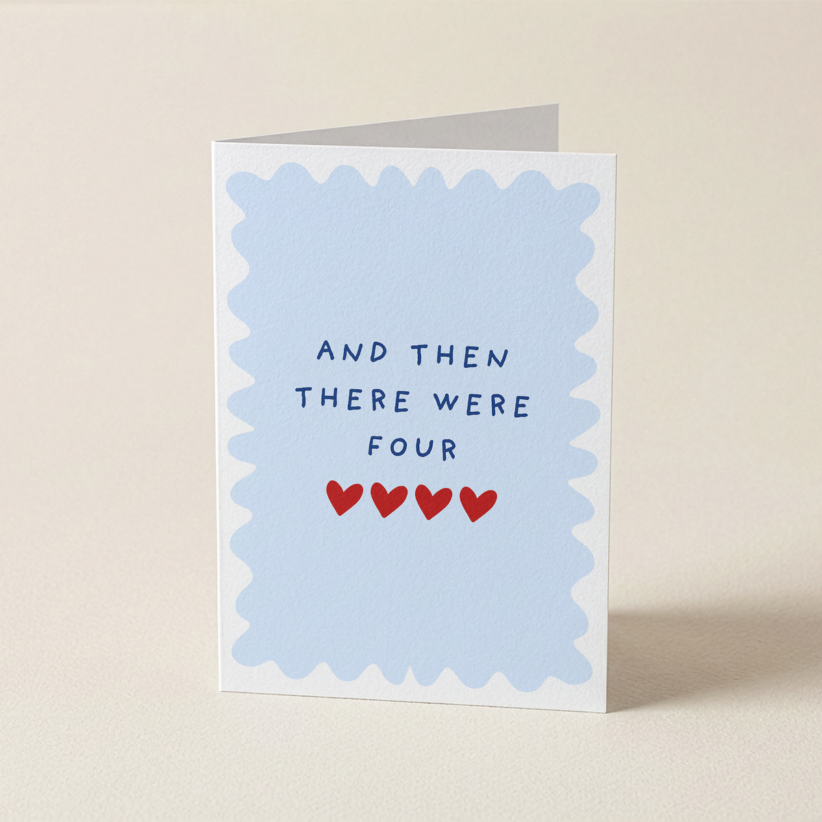 A folded greeting card standing upright on a light beige surface. The card has a white border with a scalloped light blue inner frame and the printed text 'AND THEN THERE WERE FOUR' above four small red hearts.