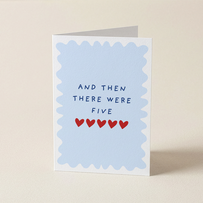 A folded greeting card standing upright on a light beige surface. The card has a white border with a scalloped light blue inner border and the printed text 'AND THEN THERE WERE FIVE' above five small red hearts.