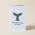 A folded greeting card standing upright on a light beige surface. The card has a pale blue front with an illustration of a whale tail decorated with Christmas lights and the text 'HAVE A WHALE-Y GOOD CHRISTMAS!'.