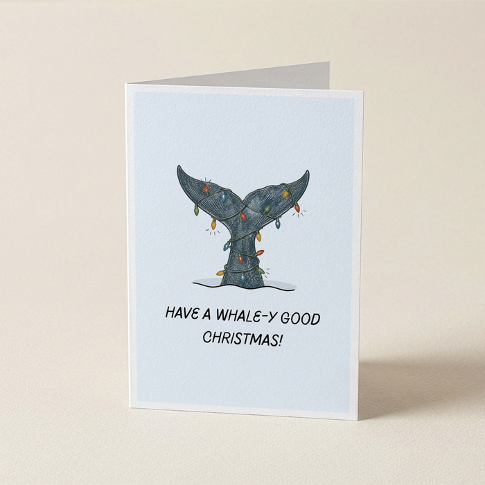 A folded greeting card standing upright on a light beige surface. The card has a pale blue front with an illustration of a whale tail decorated with Christmas lights and the text 'HAVE A WHALE-Y GOOD CHRISTMAS!'.