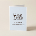 A folded greeting card standing upright on a light beige surface. The card has a pale blue front with a simple line drawing of a wine glass and a cheese board, plus the text 'TIS THE SEASON! (FOR EATING CHEESE AND DRINKING WINE.