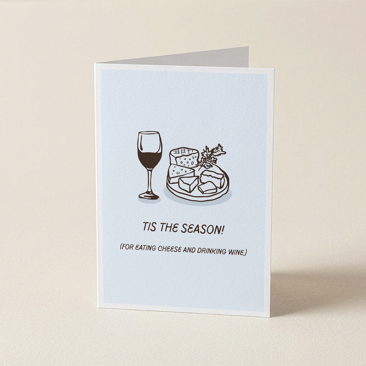A folded greeting card standing upright on a light beige surface. The card has a pale blue front with a simple line drawing of a wine glass and a cheese board, plus the text 'TIS THE SEASON! (FOR EATING CHEESE AND DRINKING WINE.