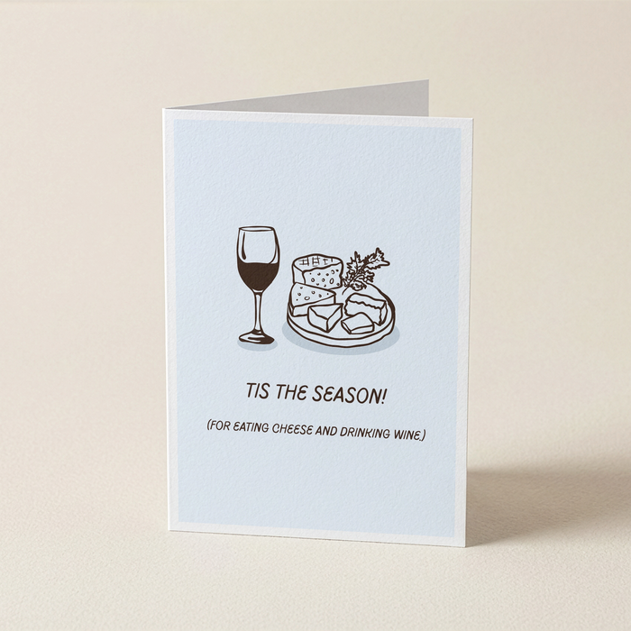 A folded greeting card standing upright on a light beige surface. The card has a pale blue front with a simple line drawing of a wine glass and a cheese board, plus the text 'TIS THE SEASON! (FOR EATING CHEESE AND DRINKING WINE.