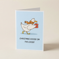 A folded greeting card standing upright on a light beige surface. The card features a white goose wearing a brown knit scarf and carrying a red gift box, with the text 'CHRISTMAS GOOSE ON THE LOOSE!' below on a pale blue background.