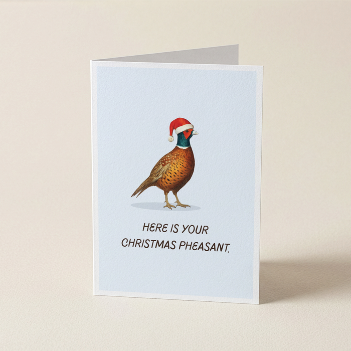 A folded greeting card standing upright on a light beige surface. The card has a pale blue center with an illustration of a pheasant wearing a red Santa hat and the text, 'HERE IS YOUR CHRISTMAS PHEASANT.