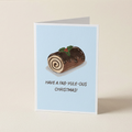 A folded greeting card standing upright on a light beige surface. The card has a pale blue background and features an illustration of a chocolate log cake with swirls, holly, and the text 'HAVE A FAB-YULE-OUS CHRISTMAS!'.