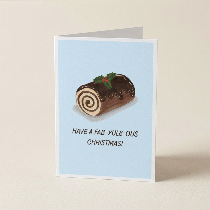 A folded greeting card standing upright on a light beige surface. The card has a pale blue background and features an illustration of a chocolate log cake with swirls, holly, and the text 'HAVE A FAB-YULE-OUS CHRISTMAS!'.
